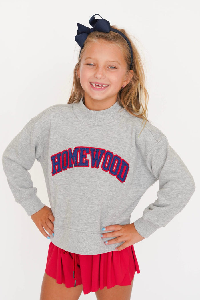 A young girl smiling and posing in a heather gray crewneck sweatshirt with 'HOMEWOOD' printed in red and blue arched lettering across the chest, paired with a red pleated skirt and a navy blue hair bow.