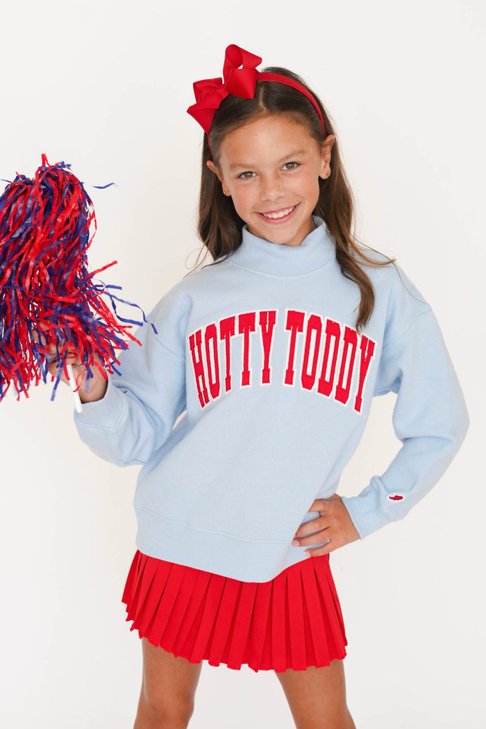 A young girl cheerleader wearing a light blue sweatshirt with 'HOTTY TODDY' printed in red across the chest, a red pleated skirt, and holding a red and blue pom-pom. She is posing against a clean white background.