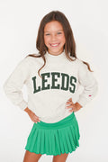 A young girl smiling and posing in a white mock-neck sweatshirt with 'LEEDS' printed in dark green collegiate letters, paired with a bright green pleated tennis skirt, against a plain white background.