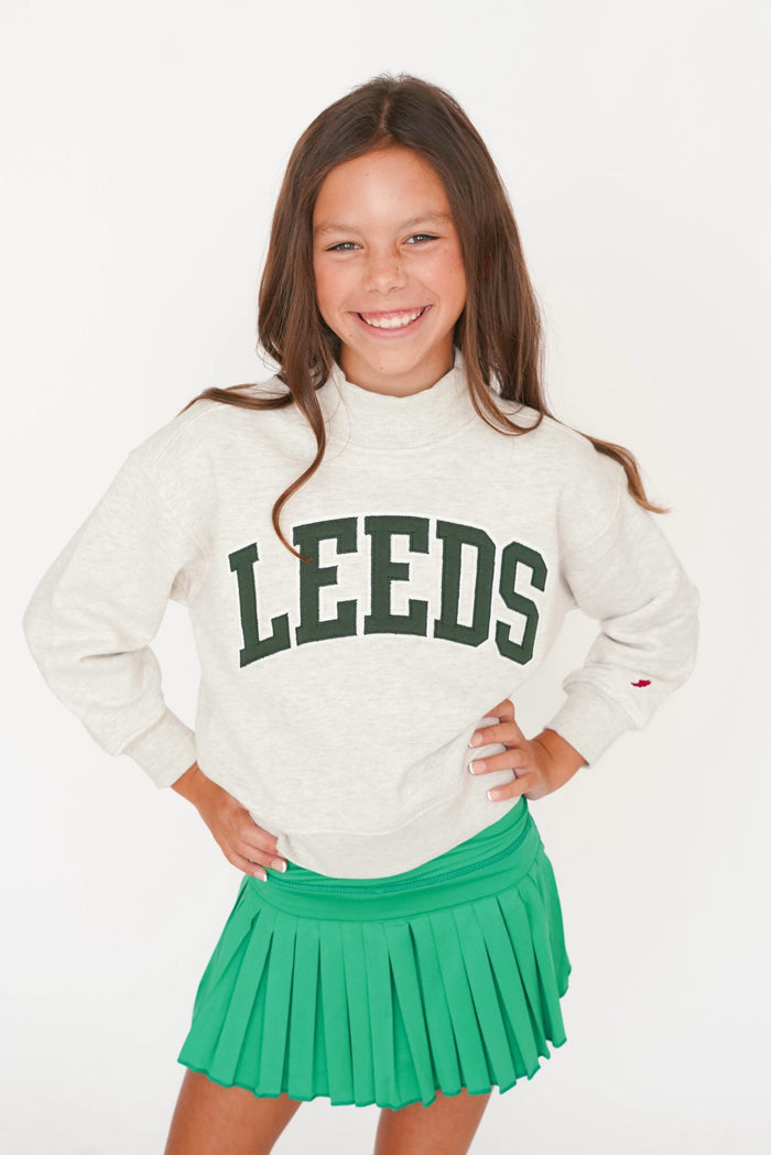A young girl smiling and posing in a white mock-neck sweatshirt with 'LEEDS' printed in dark green collegiate letters, paired with a bright green pleated tennis skirt, against a plain white background.