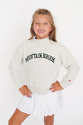 A young girl with blonde hair smiling, wearing a light heather gray sweatshirt with 'MOUNTAIN BROOK' printed in black arched letters across the chest, paired with a white pleated tennis skirt, against a plain white background.