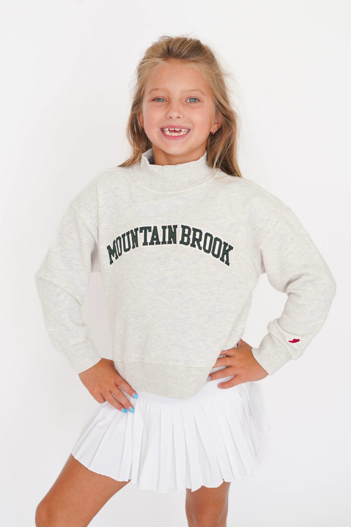 A young girl with blonde hair smiling, wearing a light heather gray sweatshirt with 'MOUNTAIN BROOK' printed in black arched letters across the chest, paired with a white pleated tennis skirt, against a plain white background.
