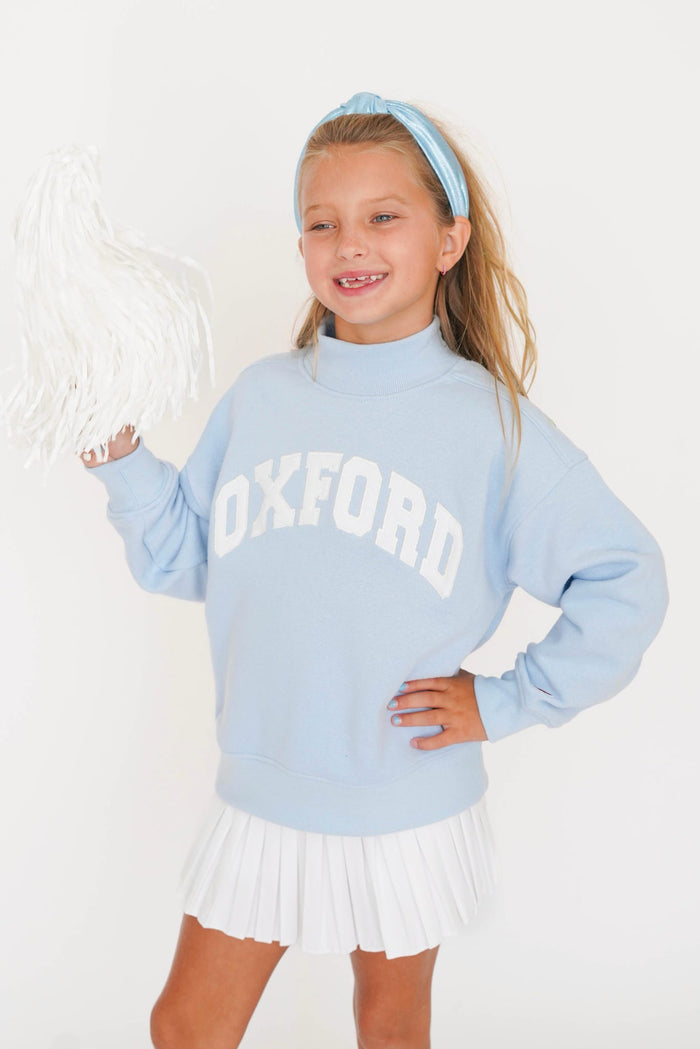 A young girl smiling and posing in a light blue mock-neck sweatshirt with 'OXFORD' printed in white arched letters across the chest, paired with a white pleated skirt and a matching light blue headband.