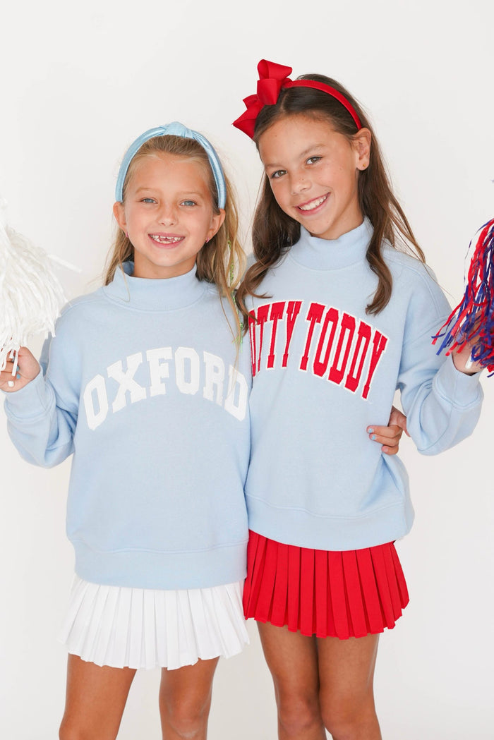 Two young girls modeling light blue sweatshirts with collegiate lettering, one reading 'OXFORD' in white and the other reading 'KITTY TODDY' in red. The girl on the right also wears a red pleated skirt and a red bow headband, while the girl on the left wears a white pleated skirt and a light blue headband.