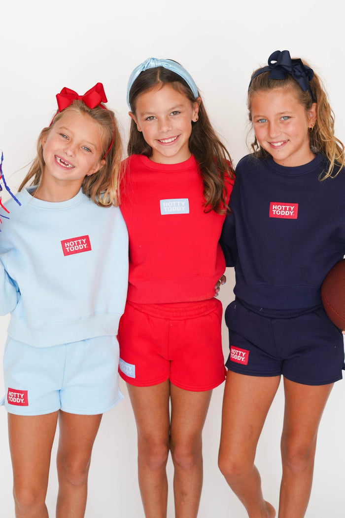 Three young girls standing together and smiling, each wearing a matching two-piece lounge set in a different color: light blue, bright red, and dark navy. Each set includes a sweatshirt and shorts with a small red square logo on the chest and a matching logo patch on the shorts.