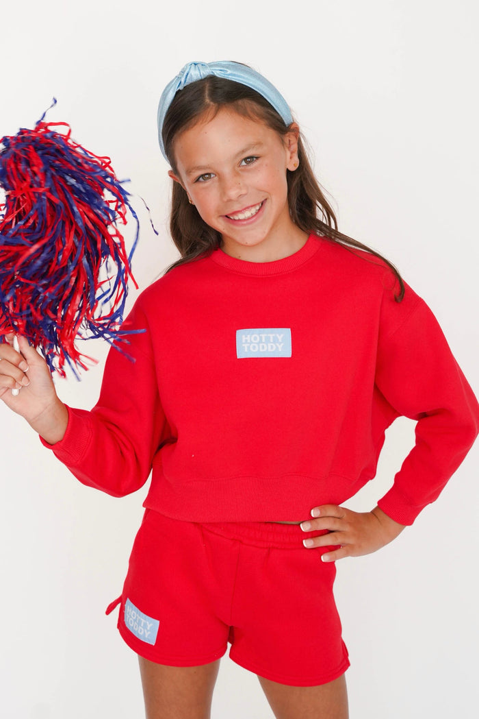 A young girl smiling and holding a red and blue pom-pom, wearing a matching red sweatshirt and shorts set with a light blue headband. The sweatshirt features a small white rectangular patch on the left chest with the text 'DO IT TODAY'.