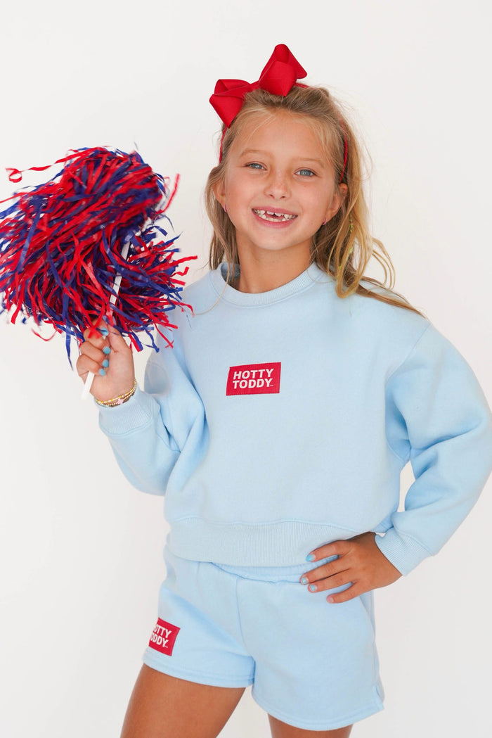 A young girl with blonde hair and a red bow is wearing a light blue sweatshirt and matching shorts set from Hotty Toddly, holding a red and blue pom-pom. The background is a clean white studio backdrop.