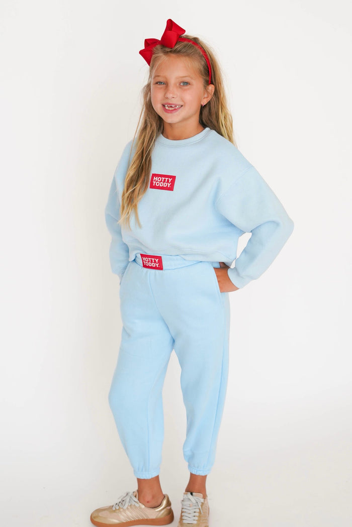 A young girl with a red bow in her hair is wearing a light blue sweatshirt and matching jogger pants with red 'TOZZY HOME' brand patches, standing against a plain white background.