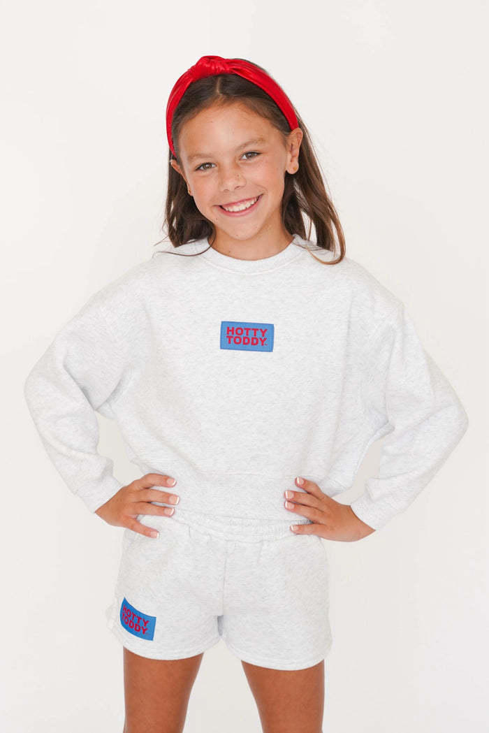 A young girl smiling and posing in a light gray heather sweatshirt and matching shorts set, featuring a red and blue 'HOTTY TODDY' logo patch on the chest and thigh. She is wearing a red headband against a clean white background.