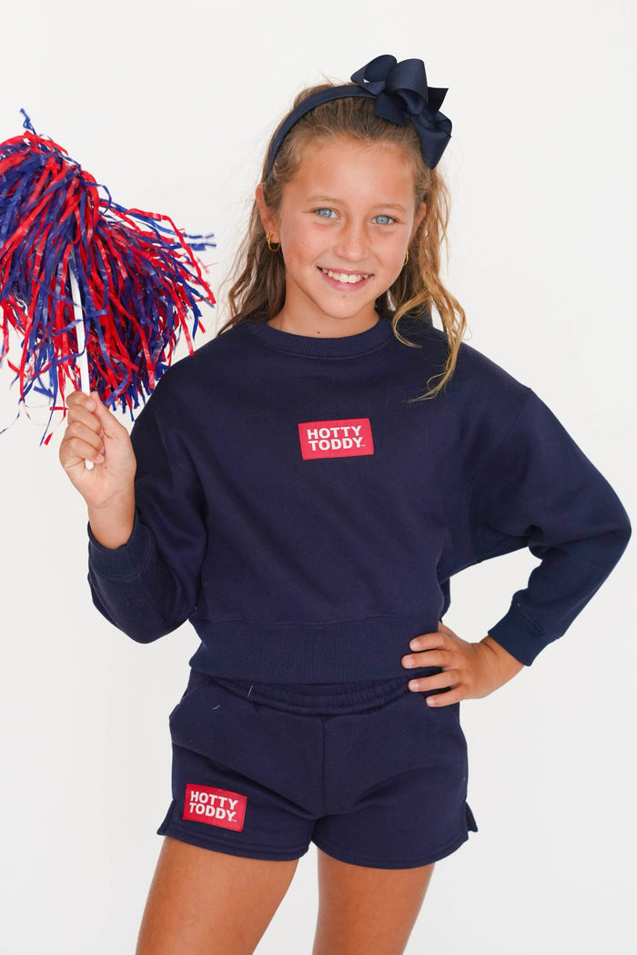 A young girl smiling and posing in a navy blue sweatshirt and matching shorts set, both featuring a red and white 'HOTTY TODAY!' logo patch on the chest and leg. She is holding a red and blue pom-pom against a clean white background.