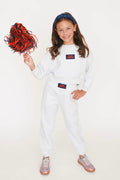 A young girl stands against a plain white studio background wearing a white sweatshirt and jogger pants set with small navy and orange logo patches. She is holding a red and navy pom-pom and wearing gray sneakers.