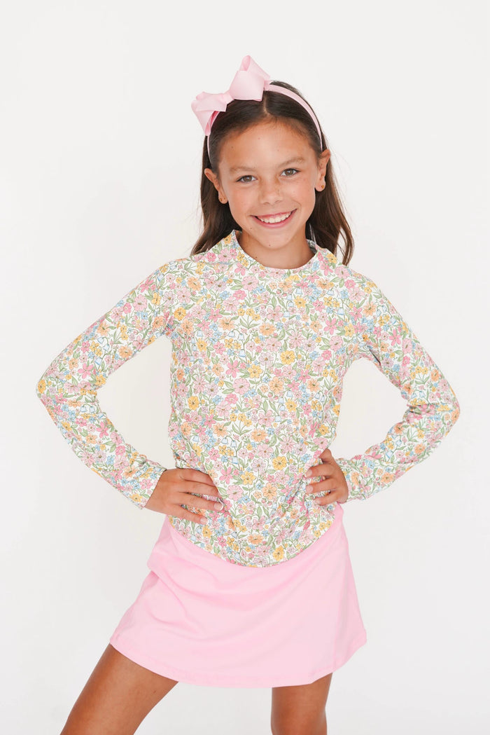 A young girl smiling and posing with her hands on her hips, wearing a long-sleeve floral print top with a pink bow headband and a pink skirt, set against a plain white background.