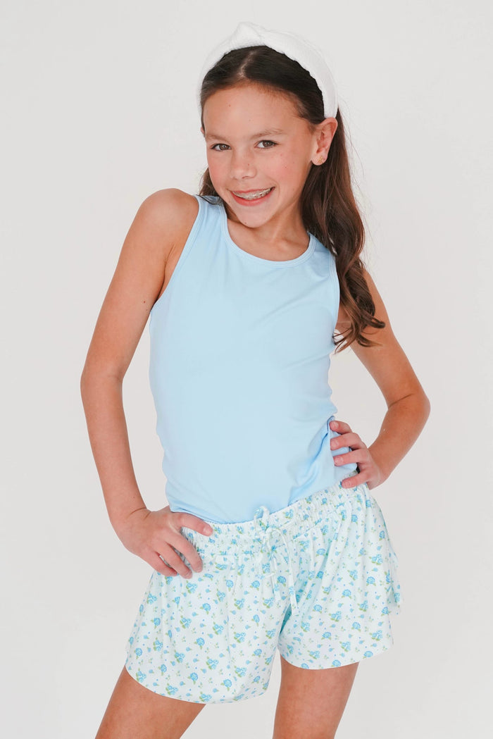 Young girl wearing a light blue sleeveless top and floral shorts on a white background