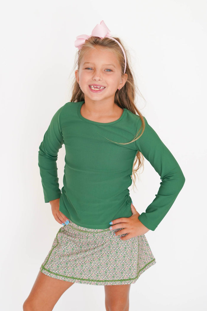 A young girl with a pink bow headband smiles while posing in a green long-sleeve shirt and a green and white patterned skirt against a plain white background.