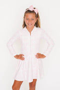 A young girl with a pink bow headband models a light pink long-sleeve zip-up dress with a subtle floral pattern against a plain white background.