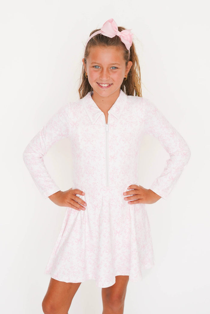 A young girl with a pink bow headband models a light pink long-sleeve zip-up dress with a subtle floral pattern against a plain white background.