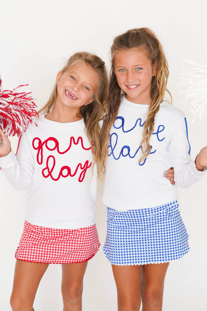 Two young girls smiling and posing together in matching white long-sleeve t-shirts with patriotic red and blue script lettering, paired with red and blue gingham skirts, against a white background.