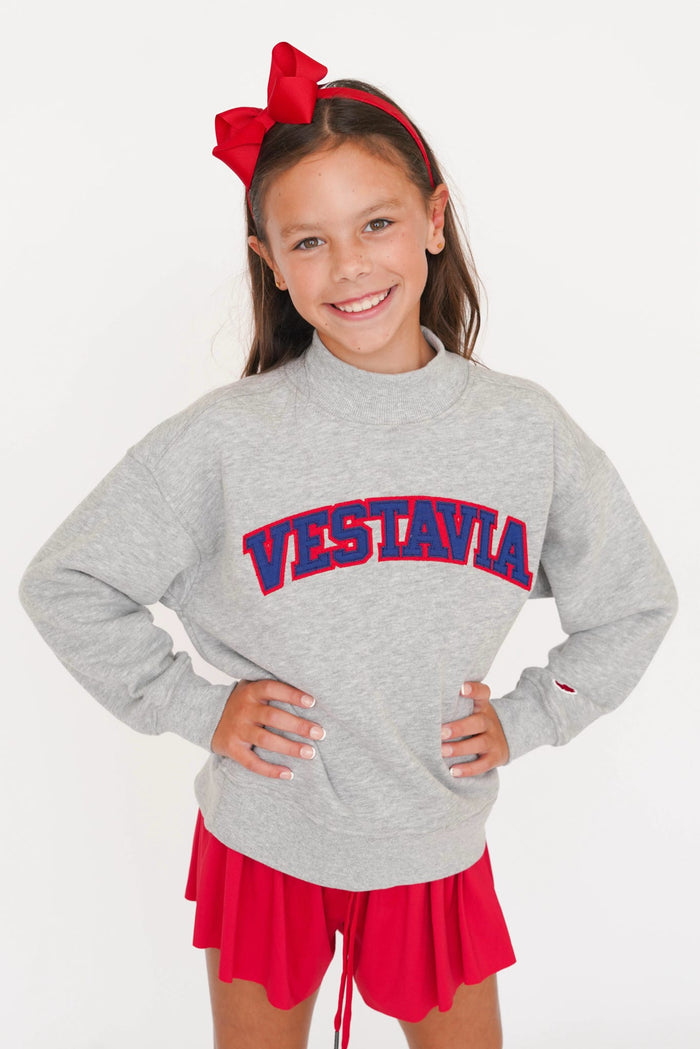 A young girl smiling and posing in a heather gray mock-neck sweatshirt with 'VESTAVIA' printed in red and blue varsity letters across the chest. She is wearing a red bow headband and a red pleated skirt against a plain white background.