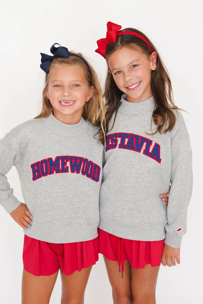 Two young girls smiling and posing together wearing matching heather gray turtleneck sweaters with 'HOMEWOOD' and 'ESTAVIA' printed in red and navy blue arched lettering, paired with red skirts.