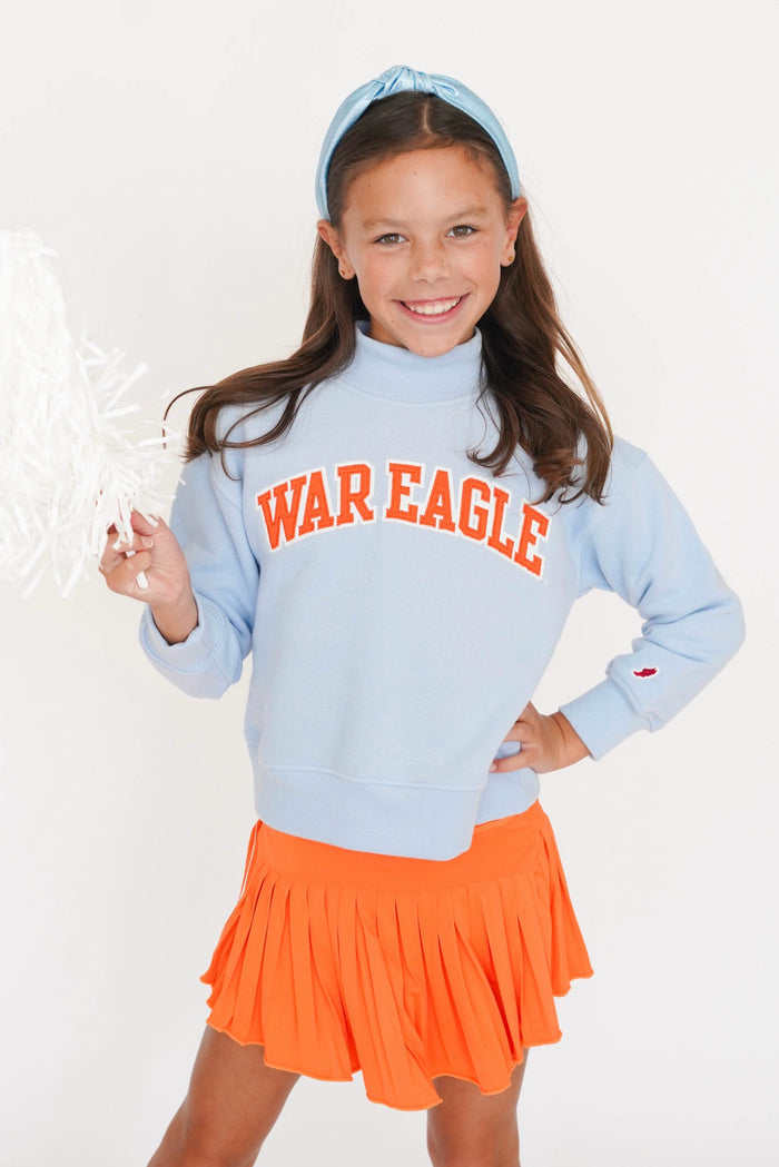 A young girl smiling and posing in a light blue cheerleader sweatshirt with 'WAR EAGLE' printed in orange, paired with an orange pleated skirt and matching blue headband, against a plain white background.