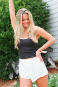 Woman in black tank top and white shorts posing outdoors with greenery in the background