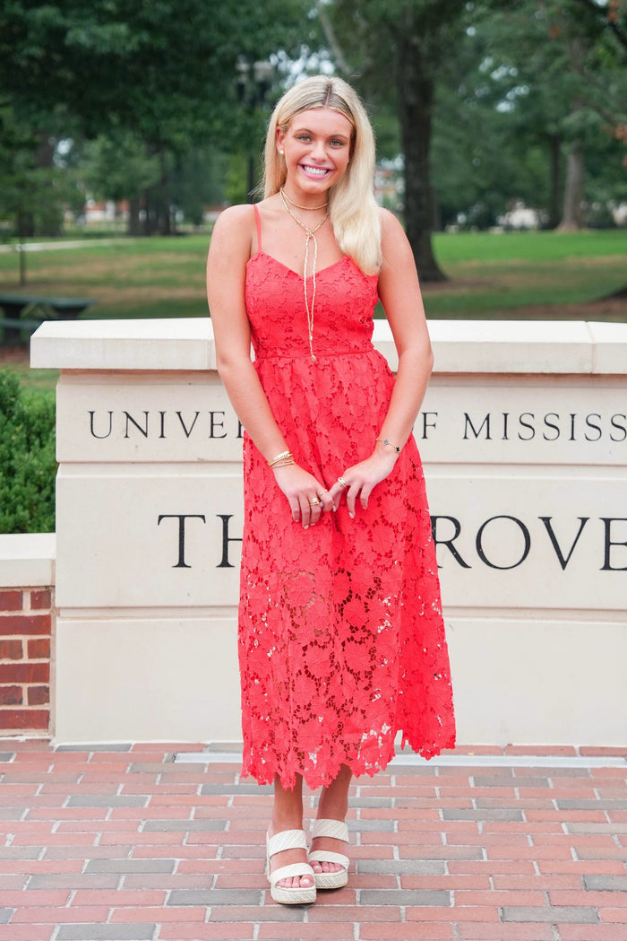 A woman stands outdoors in front of a stone sign reading University of Mississippi Throve, wearing a bright coral-red lace midi dress with thin straps and white heeled sandals.
