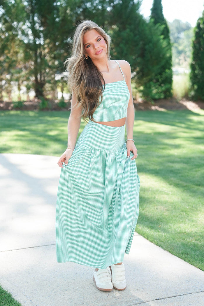Woman in a light green dress standing on a sidewalk with grass and trees in the background