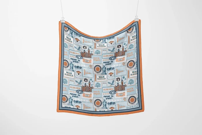 A square fabric scarf with an orange border and a light blue central print featuring vintage-style travel posters, birds, and text, hanging against a plain white wall.