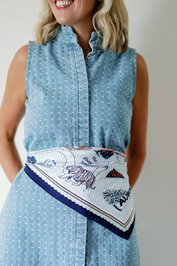 A woman wearing a sleeveless light blue patterned shirt dress with a white scarf featuring a tiger print and navy blue trim tied around her waist.