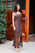 A woman wearing a brown crochet lace halter maxi dress with a tiered, ruffled hem, standing in front of a rustic wooden barn door.