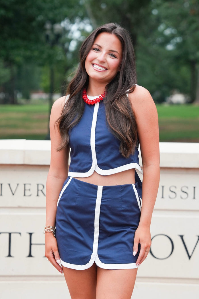 A woman smiling and wearing a navy blue two-piece athletic outfit with white piping, consisting of a sleeveless crop top and matching shorts, standing outdoors in front of a stone wall with green trees in the background.