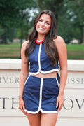 A woman smiling and wearing a navy blue two-piece athletic outfit with white piping, consisting of a sleeveless crop top and matching shorts, standing outdoors in front of a stone wall with green trees in the background.