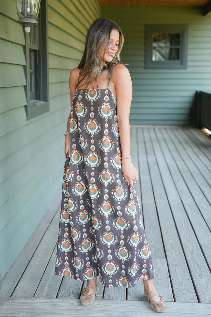 A woman wearing a sleeveless maxi dress with a brown, orange, and teal bohemian-style pattern, standing on a wooden deck in front of a house with green siding.