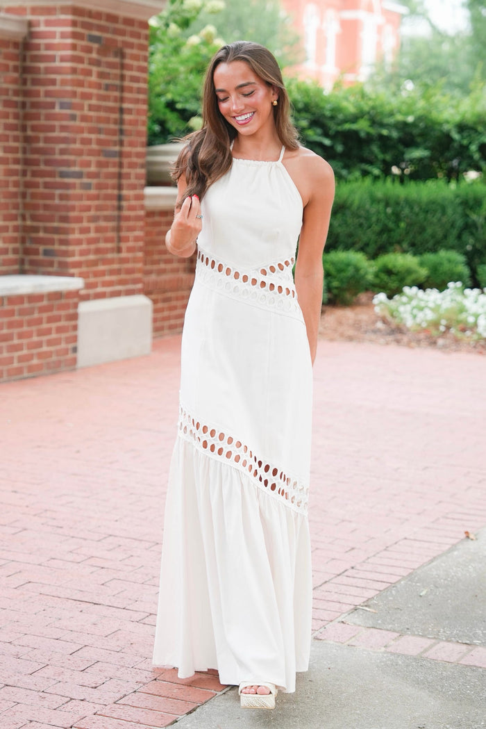 A woman wearing a sleeveless white halter maxi dress with eyelet cutout details at the waist and hem, standing outdoors on a brick walkway.