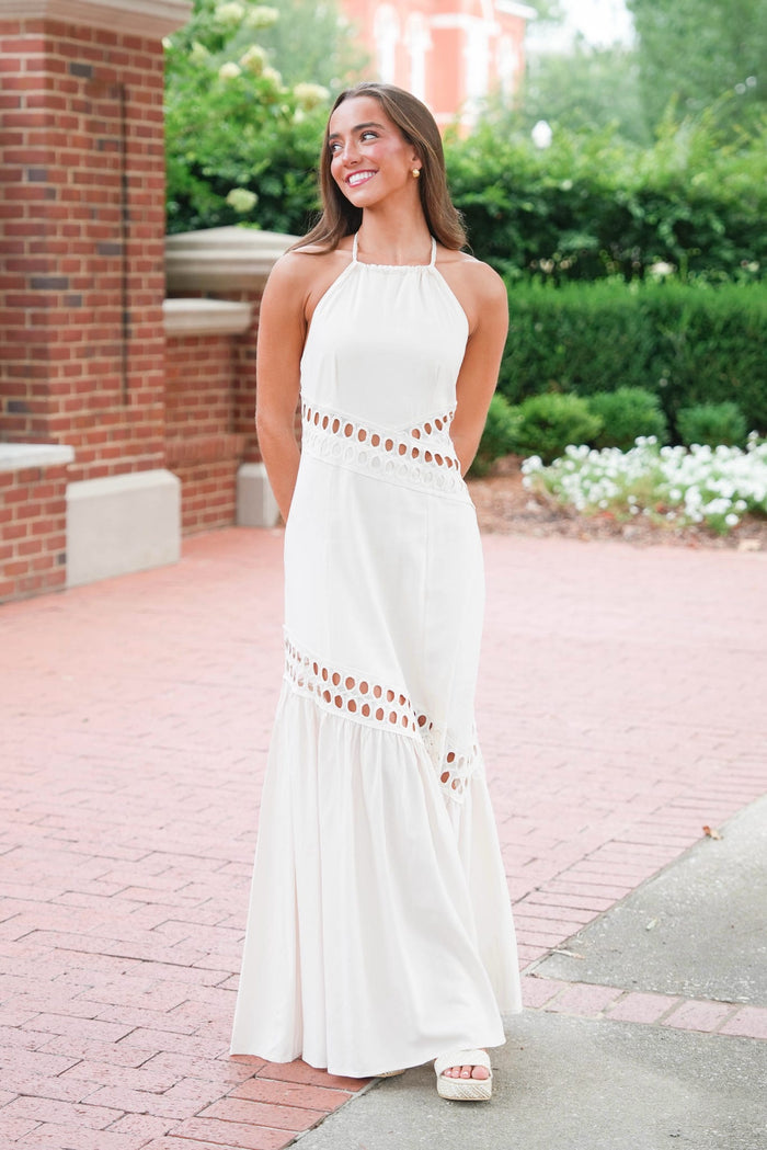 A woman wearing a white halter-neck maxi dress with cutout details at the waist and hem, standing outdoors on a brick walkway with greenery and a brick building in the background.