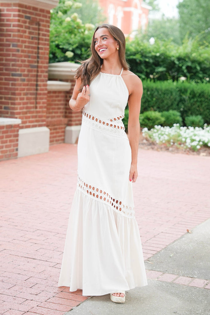 A woman wearing a sleeveless white halter maxi dress with cutout details at the waist and hem, standing outdoors on a brick walkway with greenery and a brick building in the background.