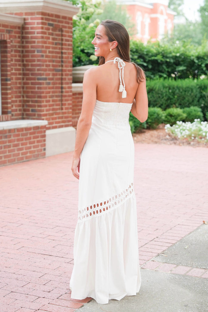 A woman wearing a strapless white maxi dress with tie-back halter details and a ruffled hem, photographed outdoors on a brick walkway with greenery and a brick building in the background.
