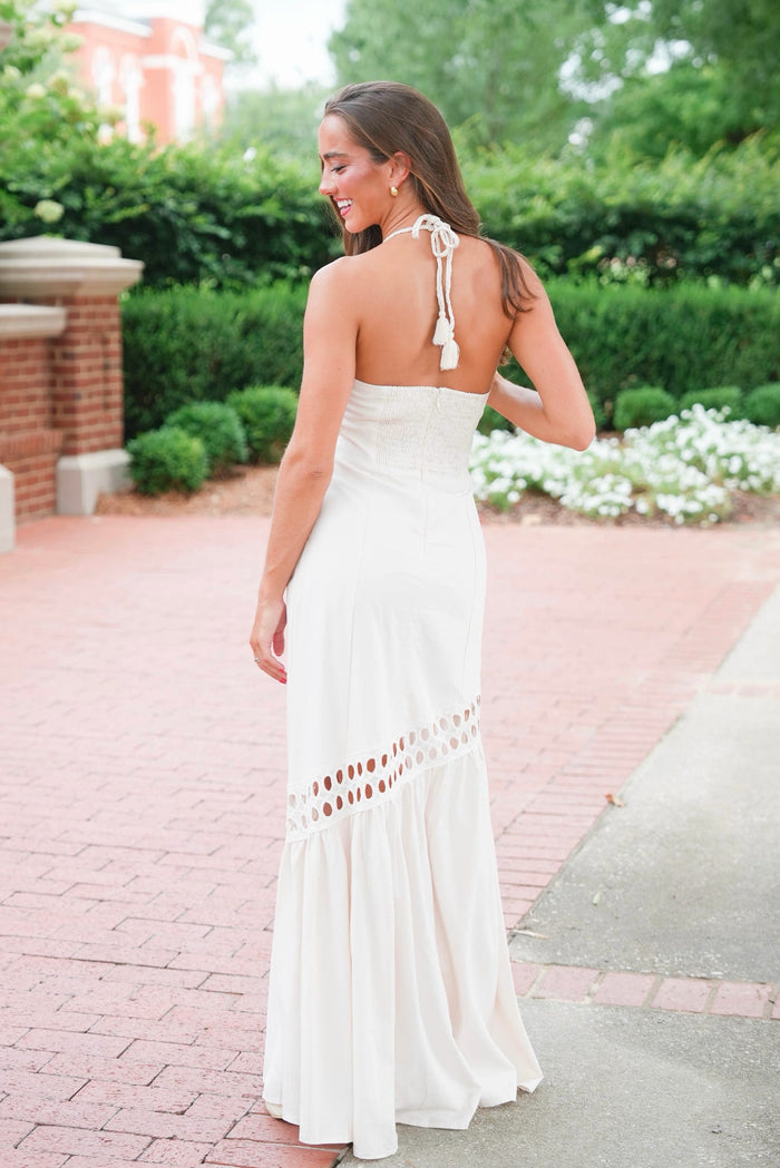 A woman wearing a sleeveless white maxi dress with tie-back straps and a ruffled hem, photographed outdoors on a brick walkway with greenery in the background.