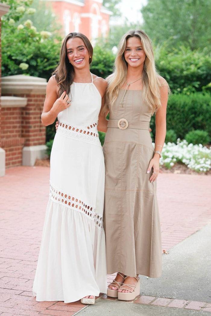 Two women standing outdoors on a brick walkway wearing matching sleeveless maxi dresses, one in white with brown cutout details and one in taupe with a large round belt buckle.