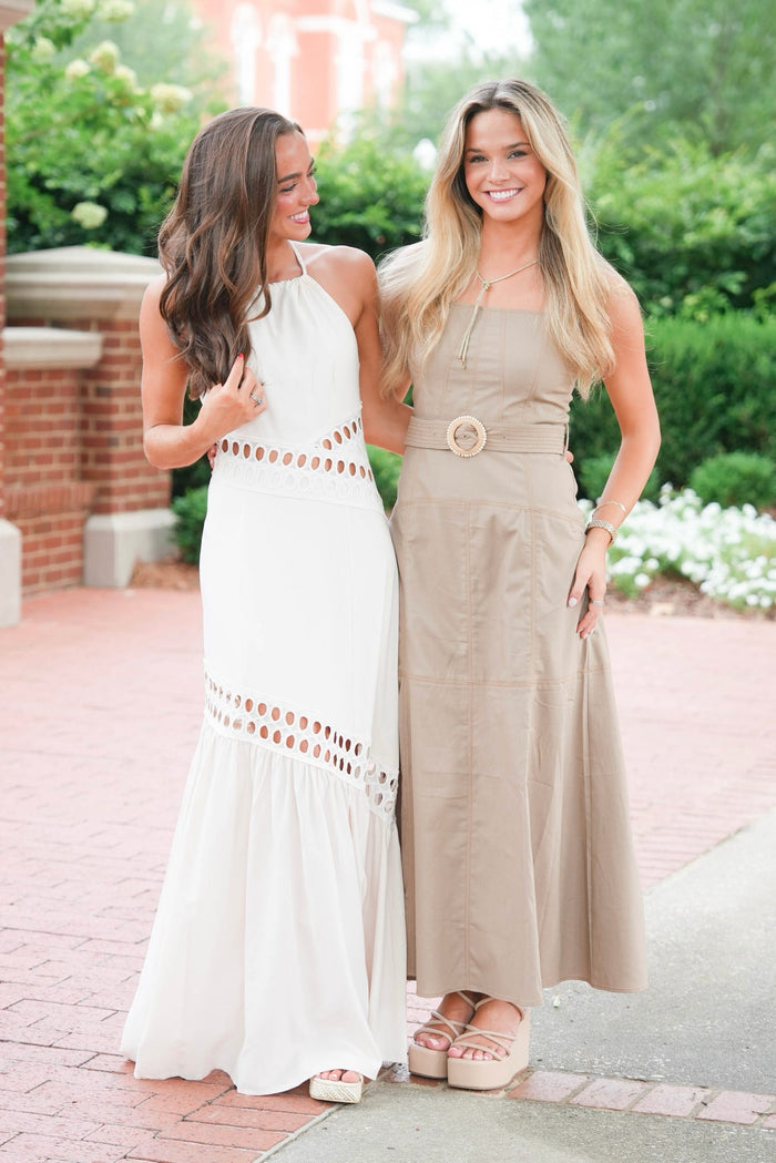 Two women standing outdoors on a brick walkway wearing long sleeveless maxi dresses, one in white with gold cutout details and one in beige with a matching belt and circular brooch.