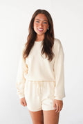 A woman modeling a matching cream-colored lounge set consisting of a long-sleeve sweatshirt and drawstring shorts, set against a plain white studio background.