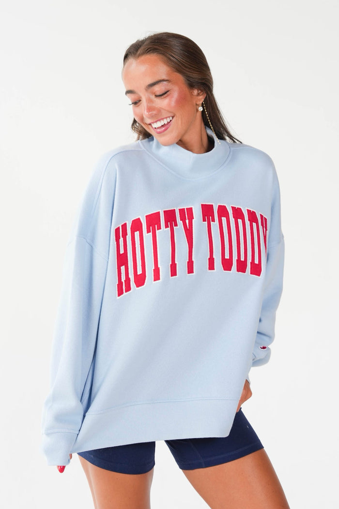 A woman wearing a light blue sweatshirt with 'HOTTY TODDY' printed in red across the chest, paired with navy blue shorts, against a plain white background.