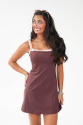 A woman modeling a sleeveless mauve-brown mini dress with light pink ruffled trim along the scoop neckline, posed against a plain white studio background.