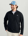 A man wearing a dark navy quarter-zip long-sleeve pullover with a small white logo on the left chest, paired with a light blue and gray trucker hat and off-white pants, against a solid light gray background.