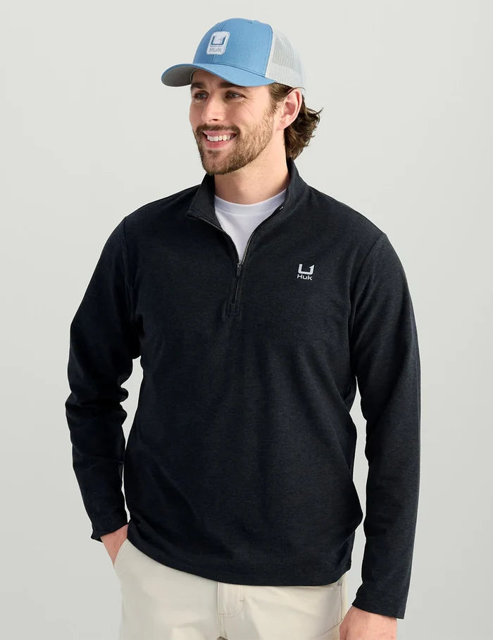 A man wearing a dark navy quarter-zip long-sleeve pullover with a small white logo on the left chest, paired with a light blue and gray trucker hat and off-white pants, against a solid light gray background.
