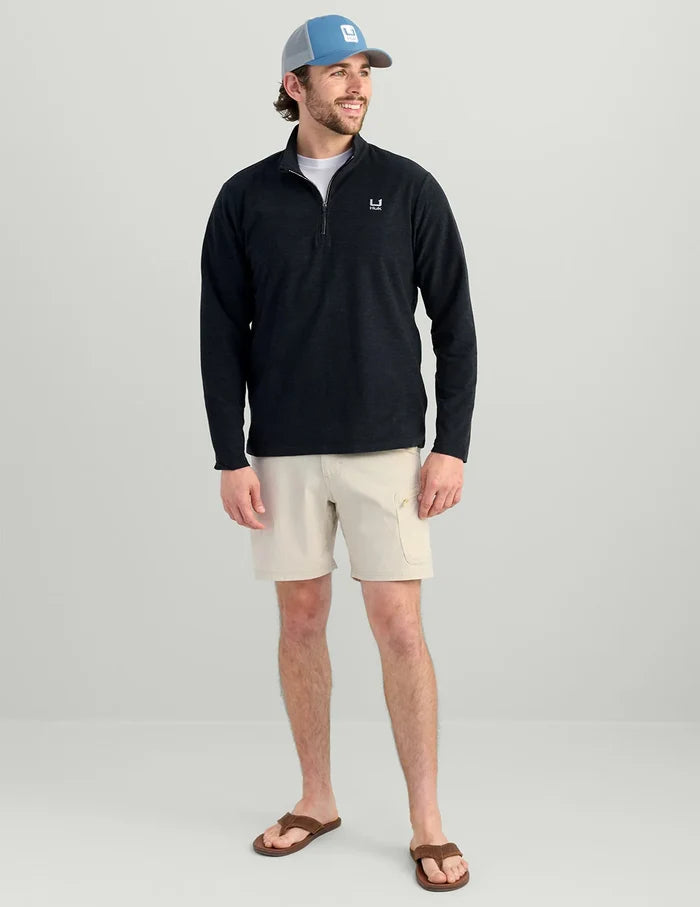 A man modeling a black long-sleeve quarter-zip pullover with a small white chest logo, paired with beige cargo shorts, brown sandals, and a blue trucker hat, against a light gray studio background.