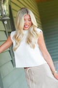 A smiling blonde woman is looking down and posing wearing a cream knitted sweater tank top paired with a beige maxi skirt. Standing, leaned in front of a green porch.