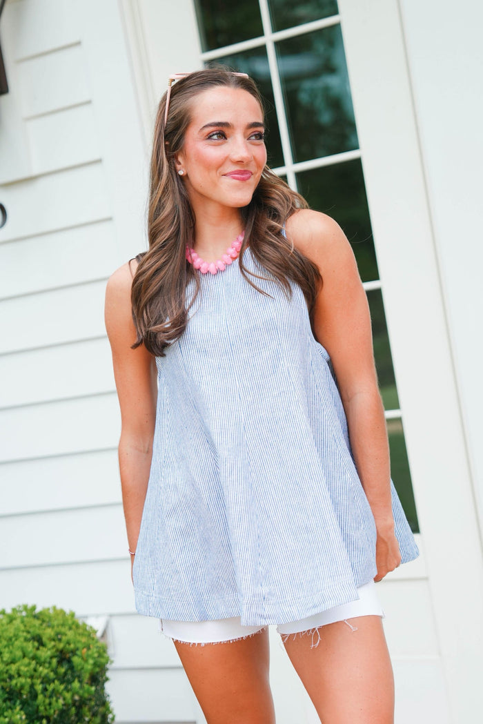 Woman wearing a light blue sleeveless top and white shorts standing in front of a white house.