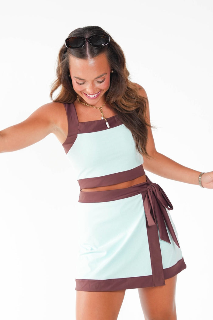 A woman modeling a two-piece sleeveless set featuring a light mint green top and matching skirt with contrasting dark brown trim, against a plain white background.