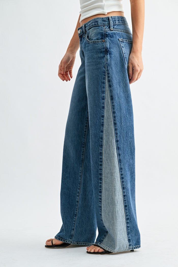 A model wearing high-waisted, wide-leg blue denim jeans with a unique dual-panel front design showing two shades of blue denim, paired with a white crop top and black sandals against a light gray studio background.